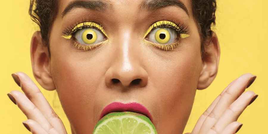 Vrouw met felgele kleurlenzen en gele eyeliner, met een schijfje limoen tussen haar lippen