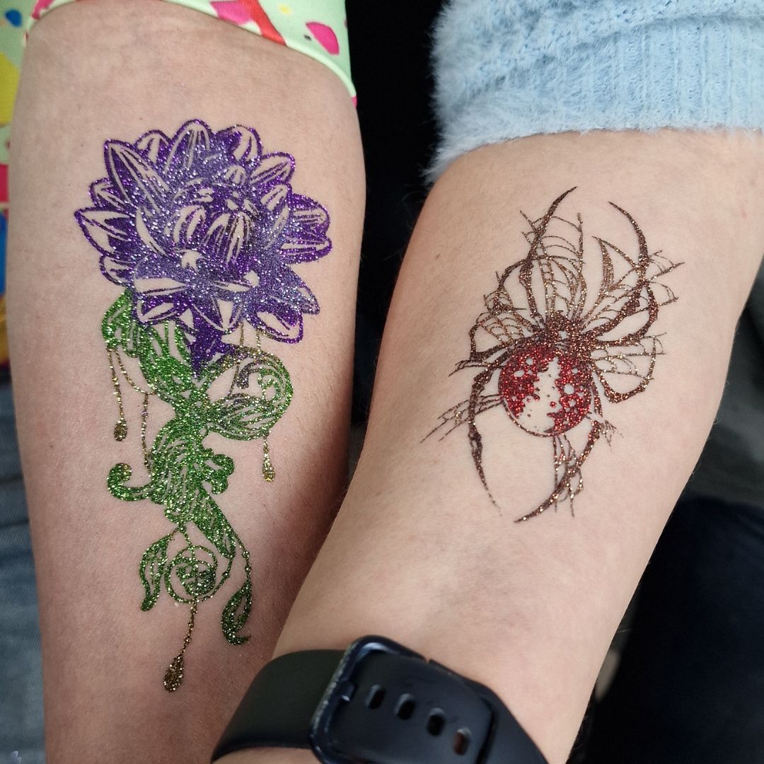 Twee glitter Quick Tattoos op de arm: een paarse bloem met groene steel en een spin met rood glitterlijf, tijdelijke tatoeages met glans, verkrijgbaar bij SchminkenGrime.nl