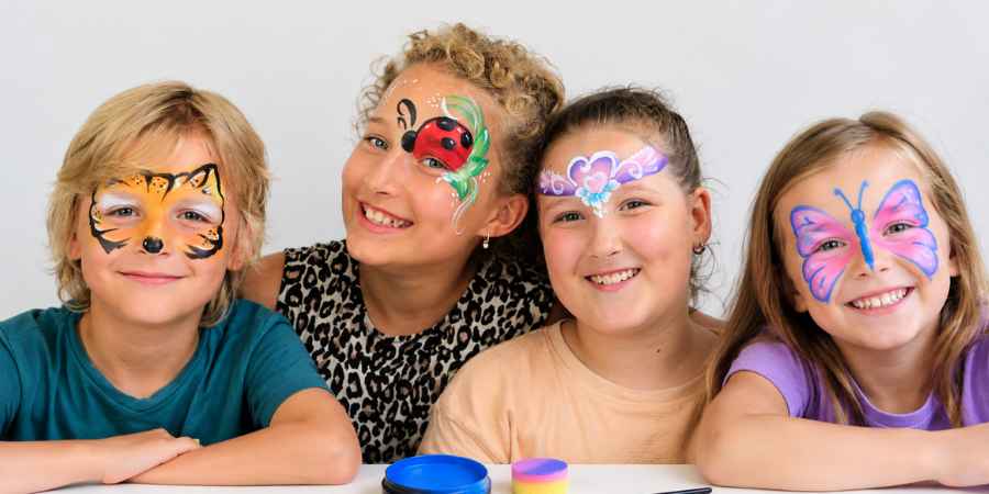 Vier lachende kinderen geschminkt als tijger, lieveheersbeestje, hartjes en vlinder, met schmink en penseel op tafel